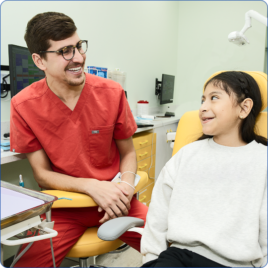 A smiling male dentist in red scrubs speaks with a young girl sitting in a yellow dental chair.