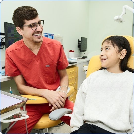 A smiling male dentist in red scrubs speaks with a young girl sitting in a yellow dental chair.